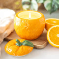 Orange shaped candle in a ceramic holder with a halved orange and decorative orange on a wooden board.