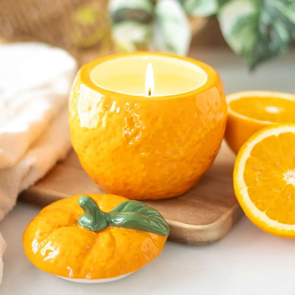 Orange shaped candle in a ceramic holder with a halved orange and decorative orange on a wooden board.