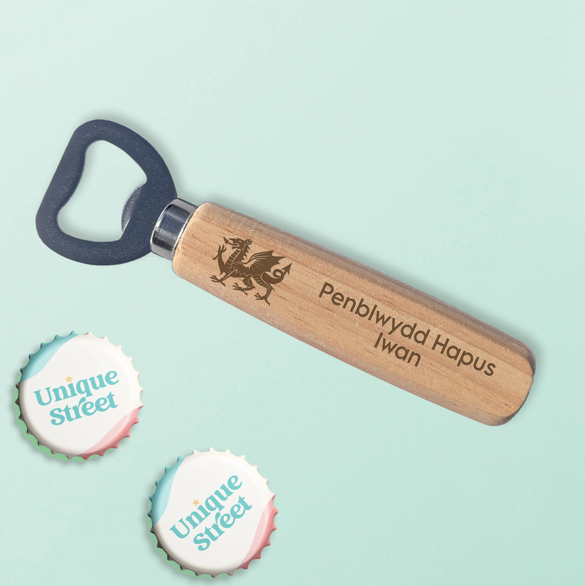 A personalized wooden bottle opener with engraved  text and Welsh dragon, shown on a pale mint background with two 'Unique Street' branded bottle cap stickers.